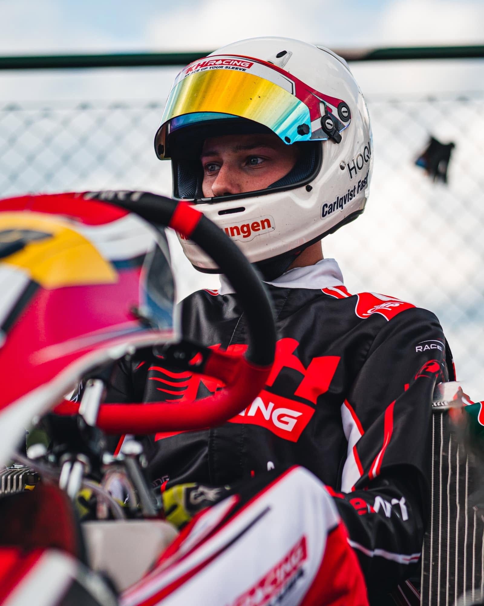 Driver in a white helmet with a KH Racing sticker and a black and red KH suit sits in the kart behind the steering wheel