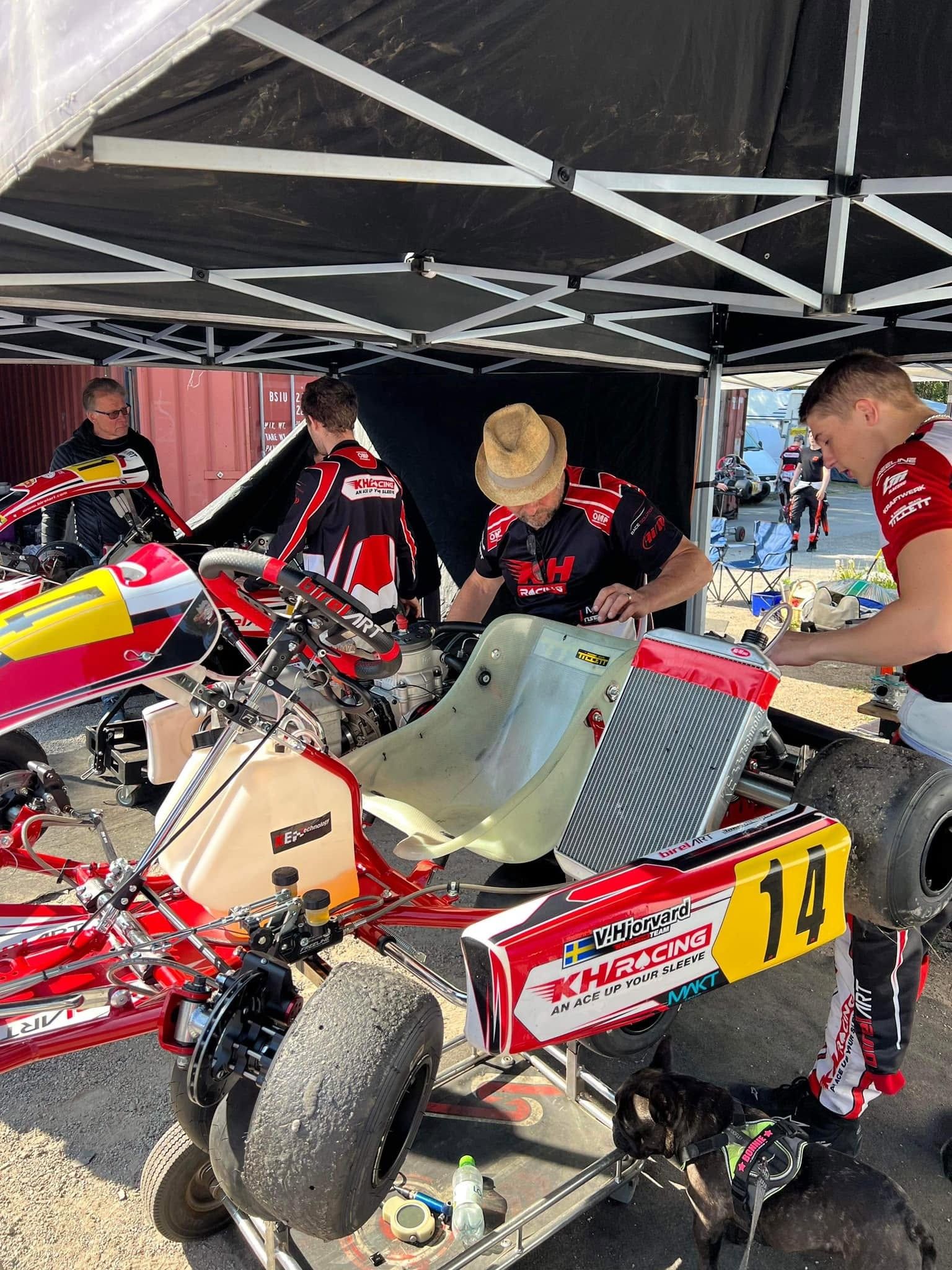 Mechanics in KH Racing kit work on KZ kart number 14, which carries a KH Racing sidepod decal, under a black canopy