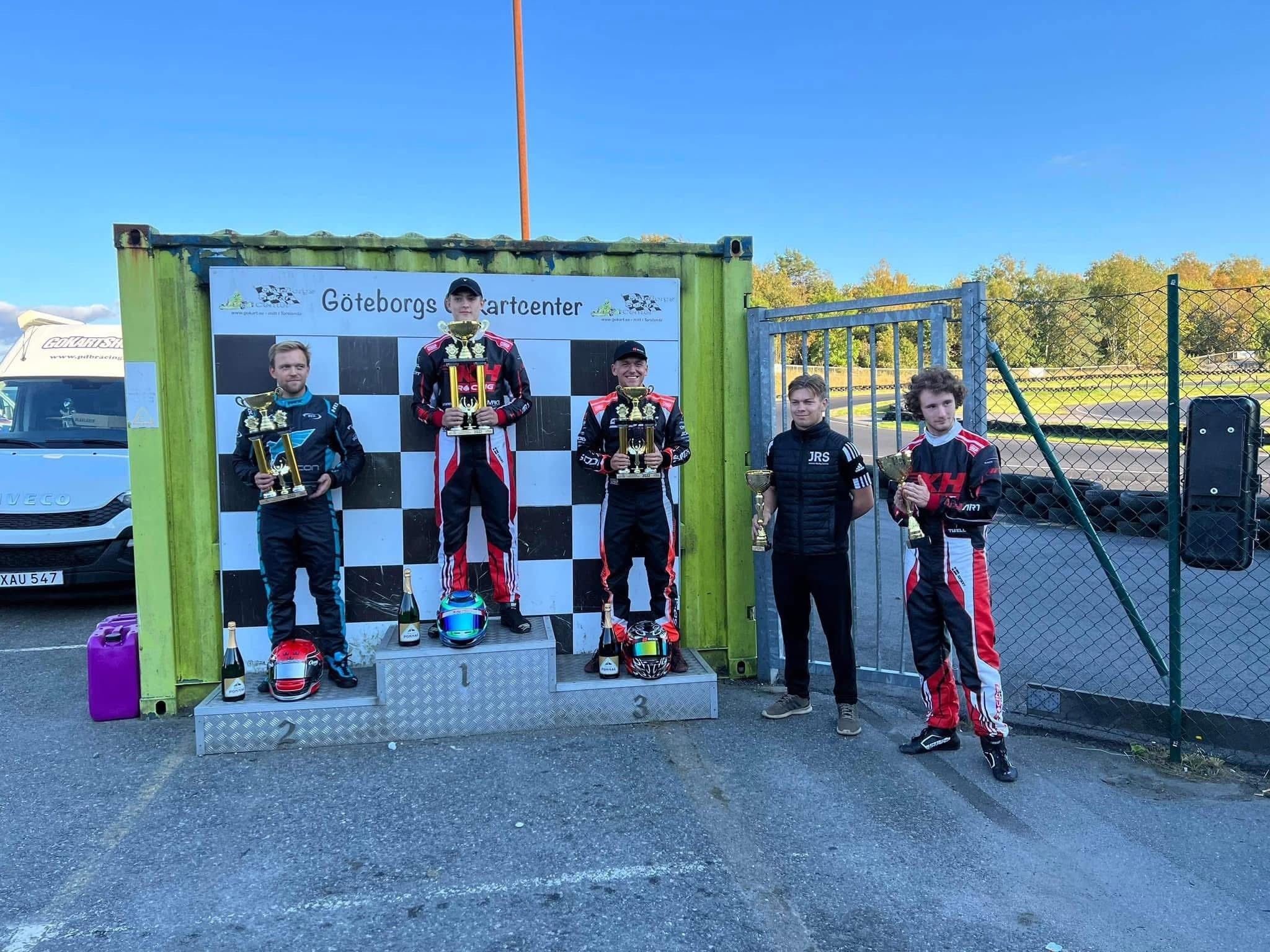 Podium at Göteborgs Kartcenter: the winner and third-placed driver in black and red KH Racing suits hold large trophies, a third KH driver stands alongside