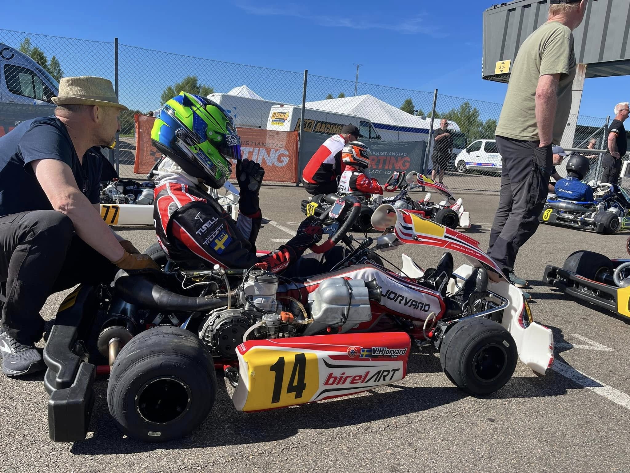 Driver in a green and blue helmet sits in Birel ART kart number 14, name V.Hjorvard on the sidepod, while a mechanic crouches alongside in the pre-grid