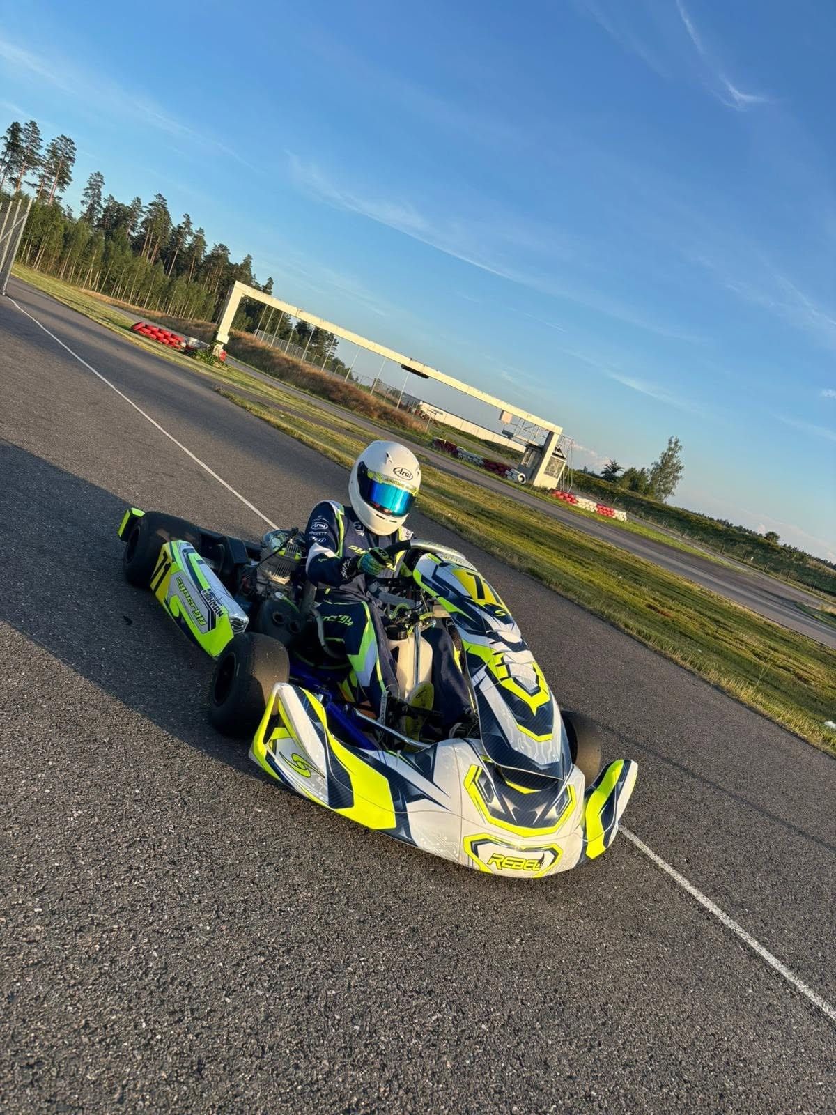 Driver in a white helmet in a Synergy Rebel kart on an empty track in evening light, a start gantry and pine forest behind