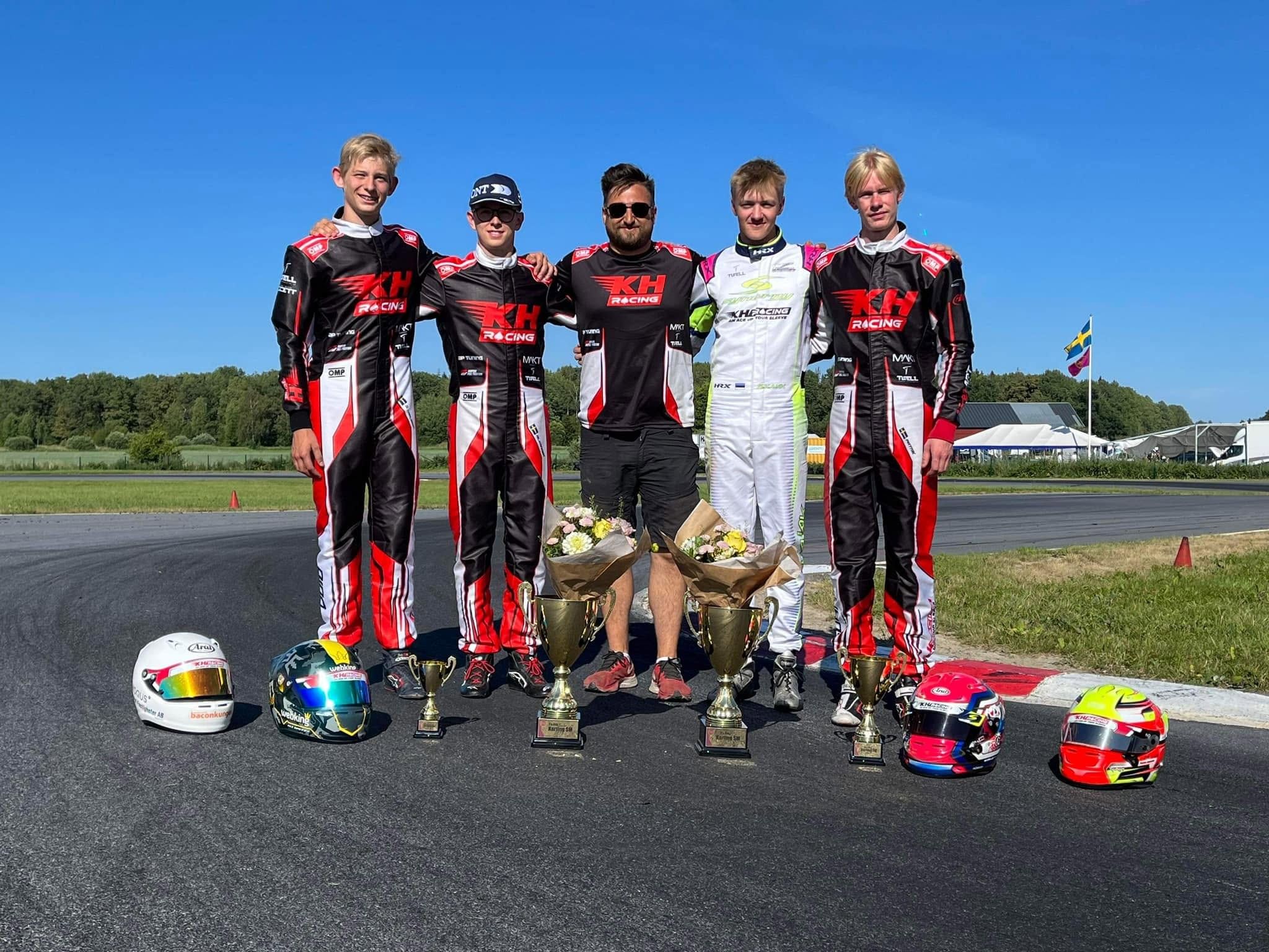 Five KH Racing members, four in black and red team suits, stand on the track with trophies, flowers and helmets in front of them