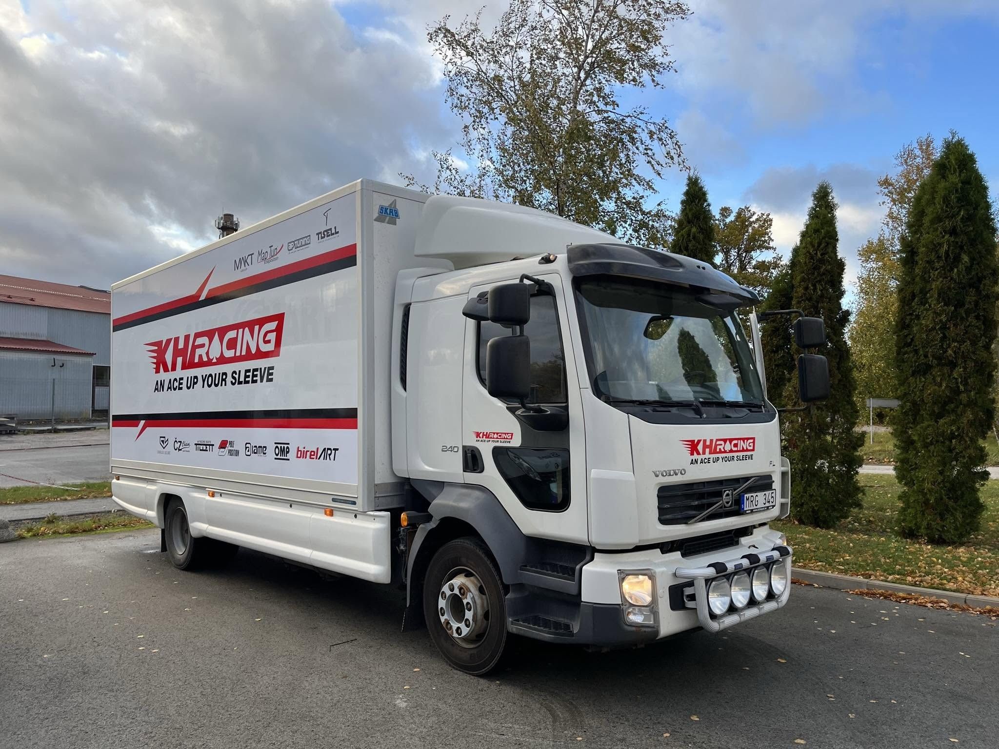 KH Racing's white truck with the team logo and sponsor decals, parked under autumn trees