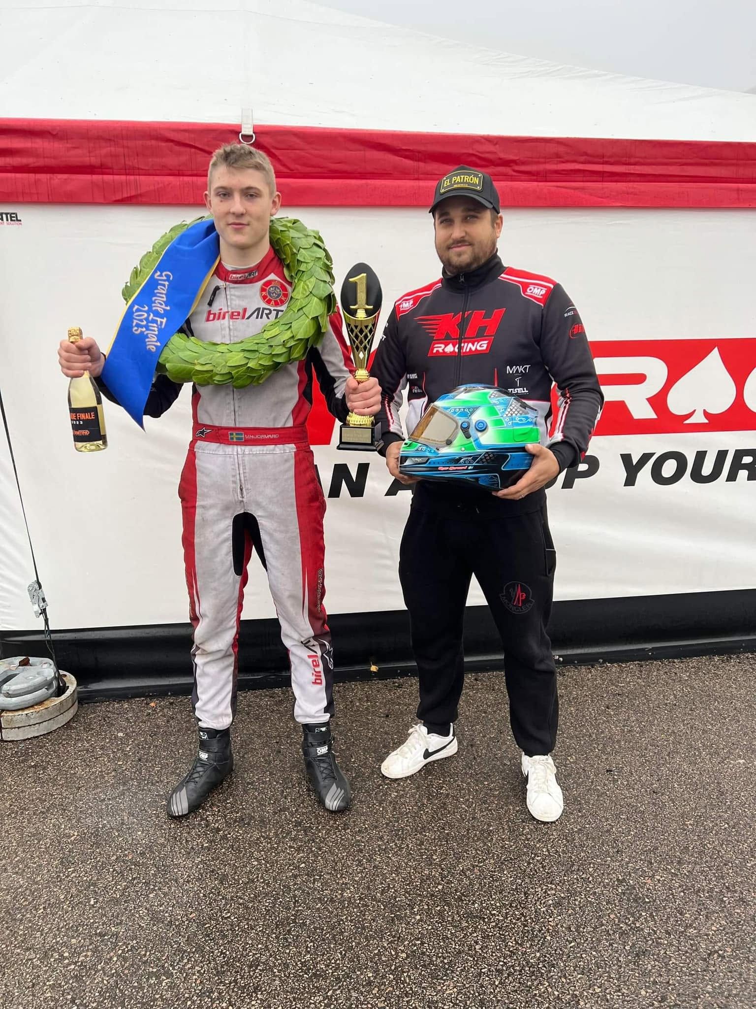 A winner with a laurel wreath and first-place trophy stands beside a team member in a KH Racing jacket in front of the KH Racing awning
