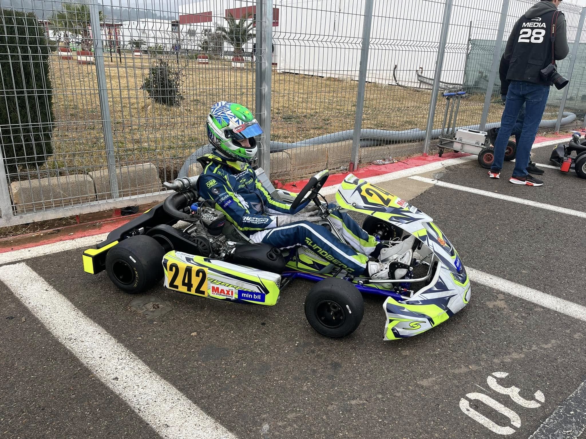 Driver in a KH Racing suit sits in Synergy kart number 242 in a paddock parking bay